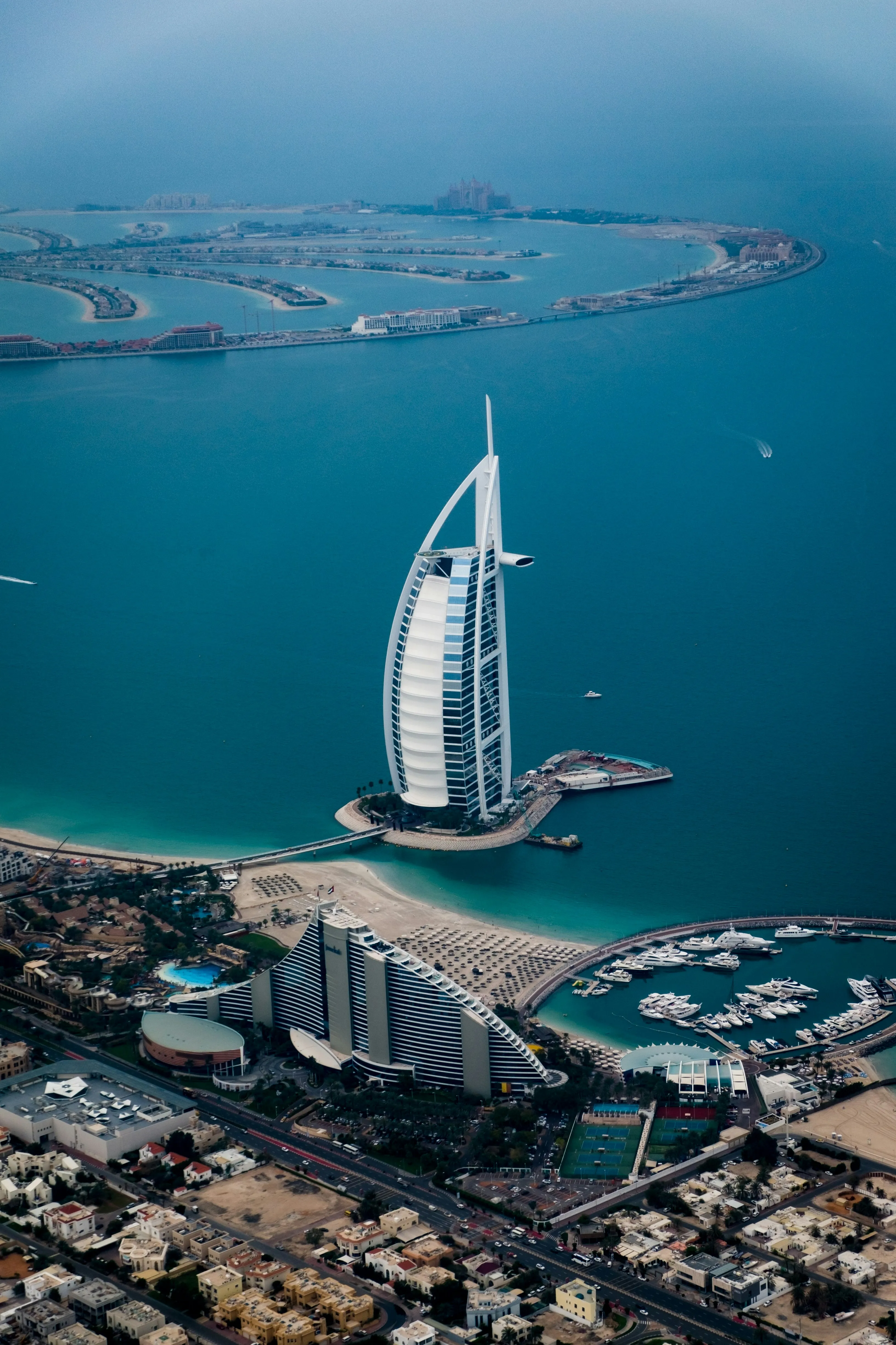 Widok z lotu ptaka na Burj Al Arab i wybrzeże Dubaju z Palm Jumeirah w tle — plaża, marina i zabudowa nadmorska.