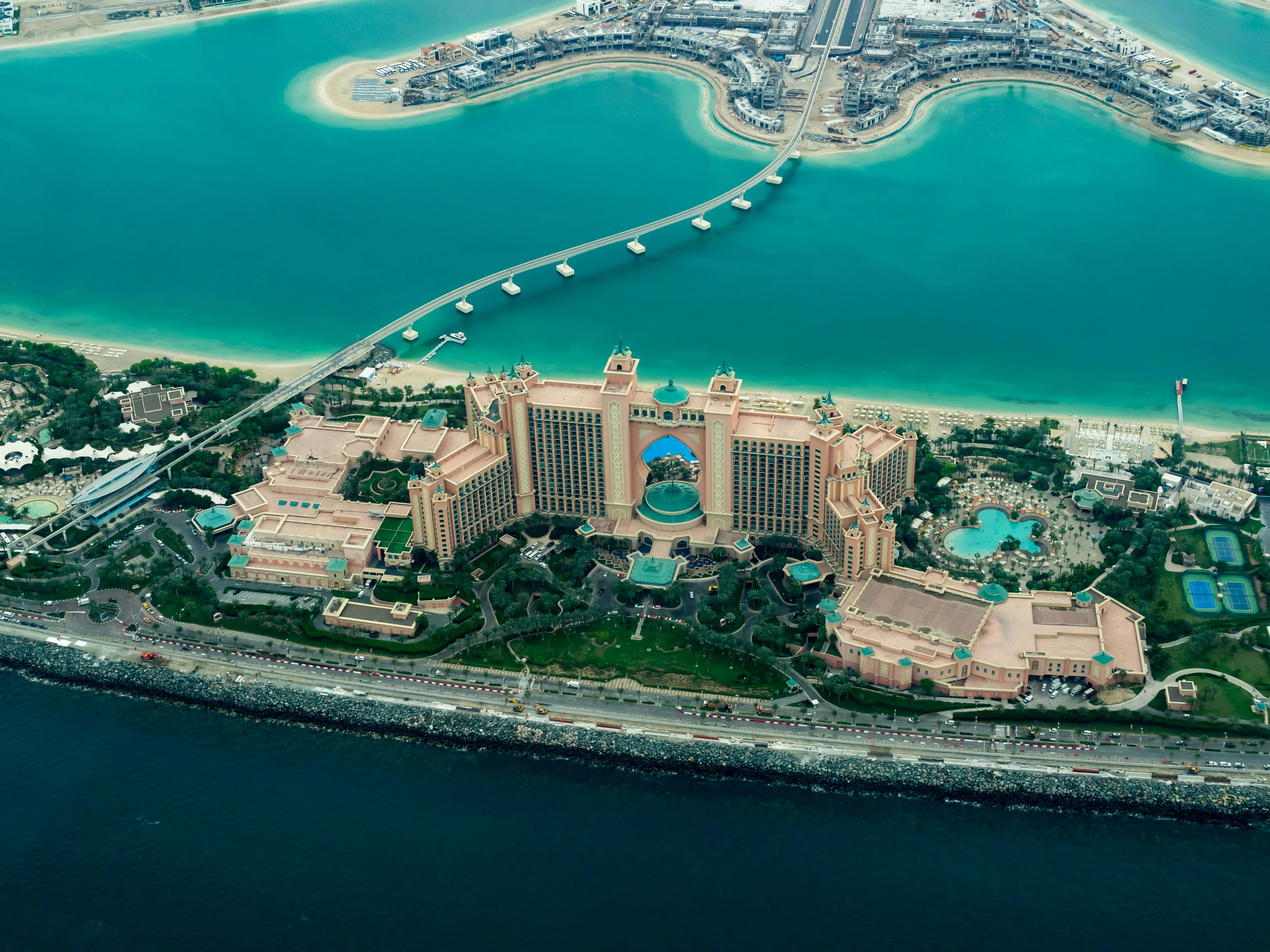 Zdjęcie lotnicze przedstawiające masywny, różowy kompleks hotelowy Atlantis The Palm na tle turkusowej wody morskiej i piaszczystej plaży. Charakterystyczny łuk w centrum budynku oraz most prowadzący w lewo na górze kadru są widoczne.